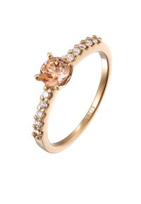 Bague en or avec un centre en pierre précieuse ronde et orange clair, entourée de petites pierres claires le long de l'anneau ; finition lisse polie.