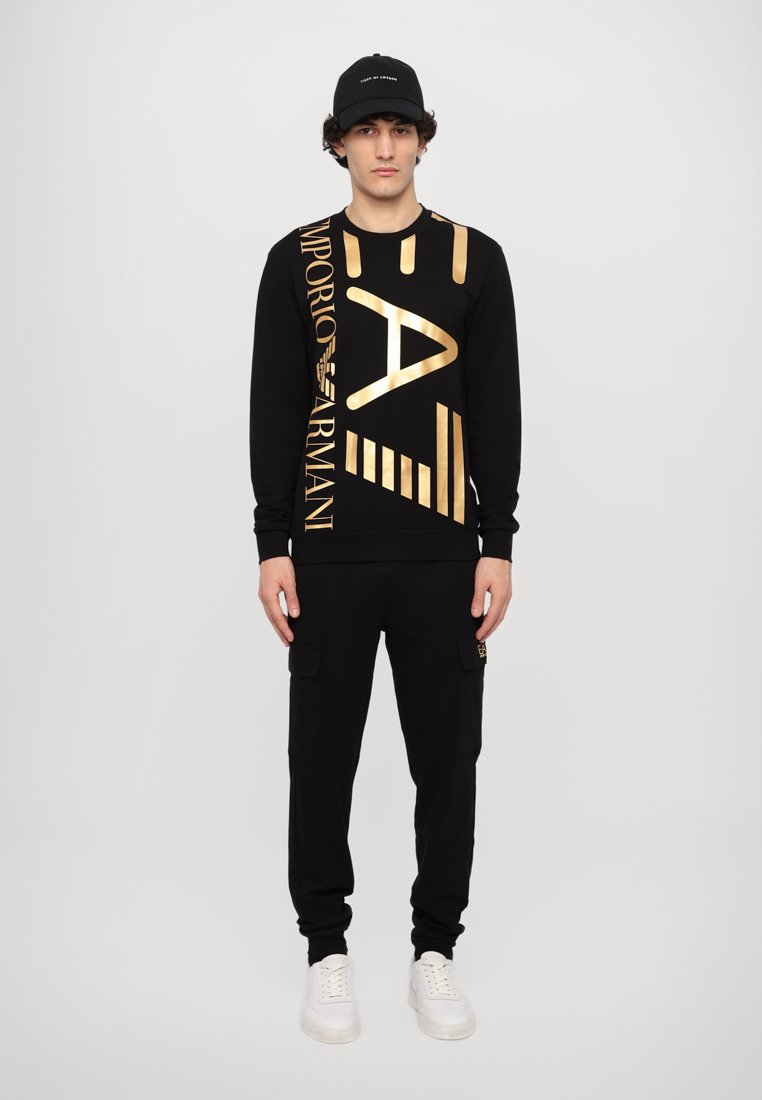 Sweatshirt noir avec des graphismes dorés, à manches longues et avec des poignets côtelés, associé à un pantalon cargo noir et des baskets blanches, porté avec une casquette noire.