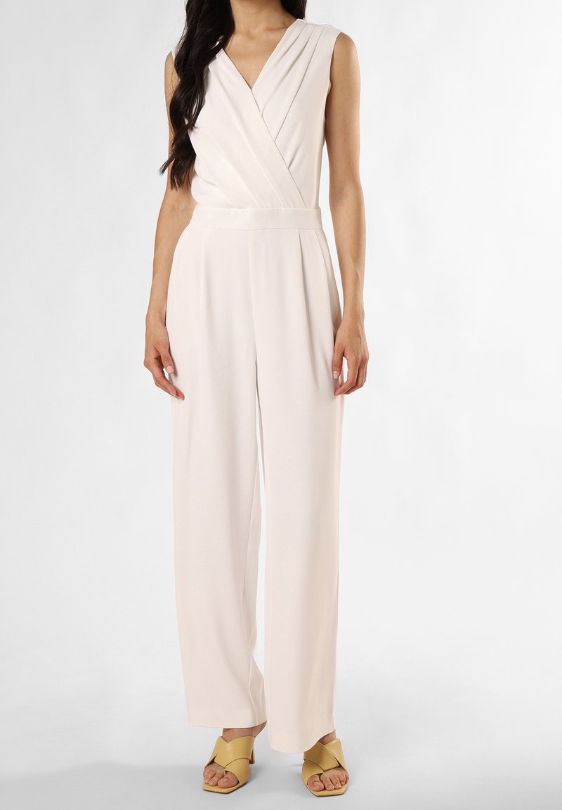 Femme portant une combinaison à bretelles blanches à encolure en V et jambes larges, avec un haut en style cache-cœur et des sandales à talons moutarde, se tenant contre un fond blanc.