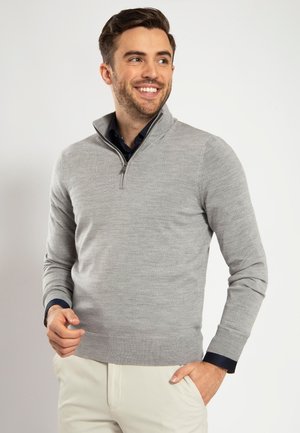 Hellgrauer Pullover mit halber Reißverschlusskragen, gerippten Bündchen und strukturiertem Strick. Getragen über einem dunklen Hemd mit Kragen, kombiniert mit cremefarbener Hose.