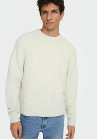 Pull clair texturé avec un col rond et des poignets côtelés, associé à un jean en denim bleu. Le tissu semble doux et chaud.