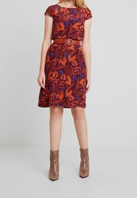 Kortärmad klänning i röd med ett blommönster av blå och orangea blad, åtdragen vid midjan med ett brunt bälte. Ankelboots med ormskinnsmönster.