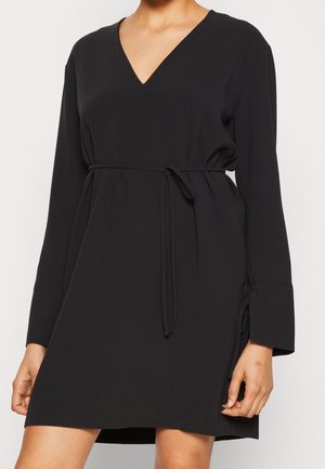 Robe noire à manches longues avec décolleté en V et fine ceinture à la taille, portée par une personne avec les mains détendues sur les côtés.