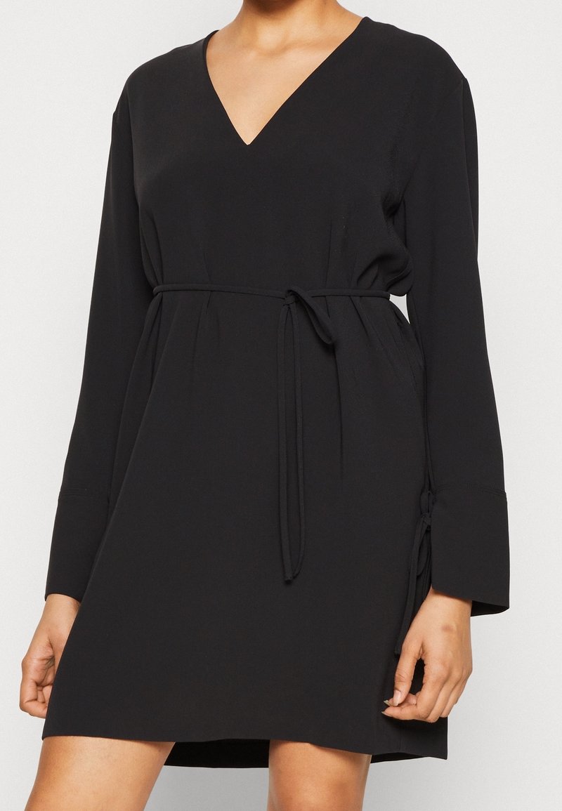 Robe noire à manches longues avec décolleté en V et fine ceinture à la taille, portée par une personne avec les mains détendues sur les côtés.