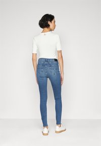 Top blanco ajustado de mangas cortas, combinado con jeans ajustados azules y zapatillas blancas con suela marrón claro. Vista trasera.