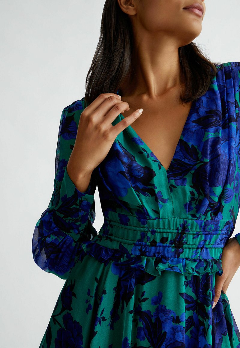 Abito verde floreale con motivi di fiori blu e neri, scollo a V, vita arricciata e maniche lunghe trasparenti con polsini arricciati.