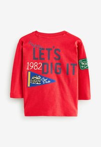 Czerwona koszulka z długim rękawem z nadrukiem "happy LET'S DIG IT 1982" oraz wyszywana niebieska chorągiewka z pojazdem i napisem "Cool Dude."