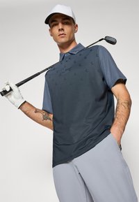Navy polo shirt with textured pattern, short sleeves, and a collar, paired with light gray pants. The model holds a golf club.