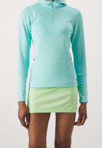 Svetle tyrkysový dlouhý pulovr s polovičním zipem a dvěma bočními kapsami, v kombinaci s mátově zelenou sukní. Hladká, elastická tkanina.