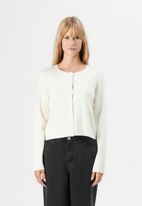 Blonde vrouw in een crèmekleurige knooptrui en hoogzitters zwarte, gestructureerde broek, staand tegen een witte achtergrond.