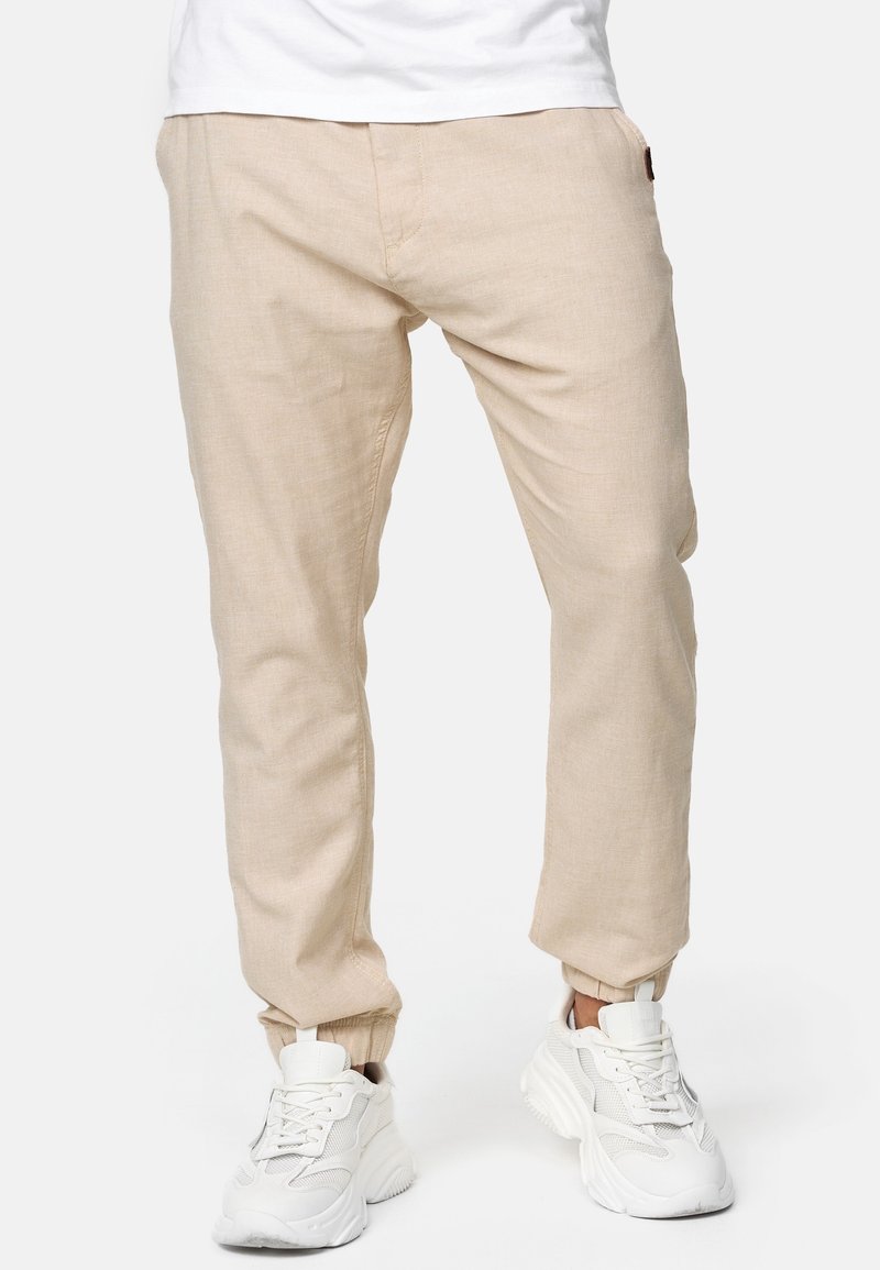 Homme portant un pantalon de jogging beige et des baskets blanches épaisses, debout les jambes légèrement écartées devant un fond uni.