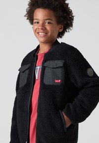 Crna sherpa jakna s sivim tkanim džepovima na prsima, rebrastim ovratnikom i Levi's logom. Ima zatvaranje na zip i teksturiranu završnicu.