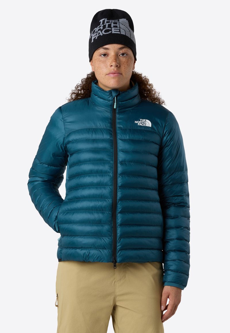Donna con capelli ricci che indossa una giacca a vento teal e un berretto nero con il logo The North Face, pantaloni beige, in piedi davanti a uno sfondo bianco.