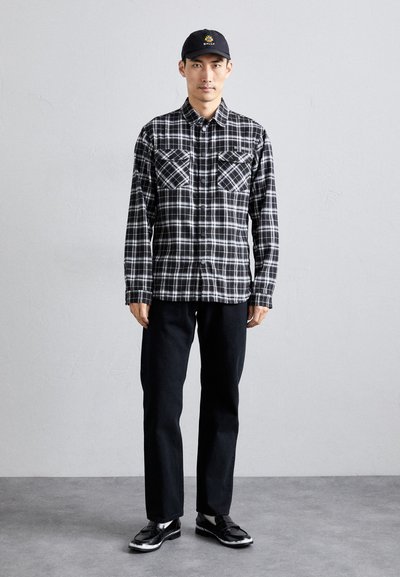 Schwarz-weiße karierte Flanellhemd mit zwei Brusttaschen, schwarze Hose und glänzende schwarze Schuhe. Die Person trägt eine schwarze Mütze mit einem Logo.