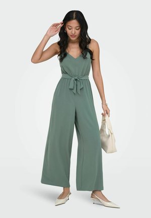 Kvinne med langt svart hår iført en ermeløs grønn jumpsuit med midjebånd, holder en beige håndveske og har på seg hvite hæler.