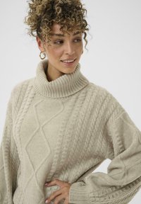 Frau mit lockigem Haar, die goldene Creolen und einen beigen, mit Kabelmuster gestrickten Rollkragenpullover trägt, lächelt und schaut zur Seite.