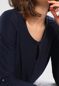 Nainen, jolla on päällään tummansininen teksturoitu pitkähihainen paita, jossa on pieni metallinen solki kaula-aukon lähellä, nojaa leukaansa käteen.