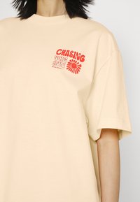 Beige bomulds T-shirt med korte ærmer. Har rød tekst "CHASING THE SUN" og et blomster graphic på venstre brystområde.