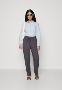 Chemise bleu clair à manches longues, pantalon gris à cordon de serrage avec coutures latérales, sandales plates noires, lunettes de soleil claires et sac à dos blanc.
