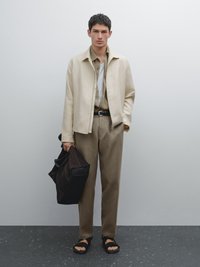 Světle béžová lněná bunda přes khaki košili a odpovídající kalhoty. Černý opasek, černé sandály a tmavá taška. Hladké textury a minimalistický design.