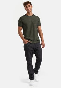 Olivgrünes T-Shirt mit kurzen Ärmeln und Brusttasche, kombiniert mit schwarzen Jeans und weißen Sneakers, das eine lockere Passform und einen strukturierten Stoff zeigt.
