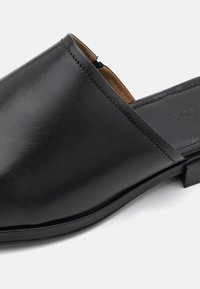Černá kožená slip-on bota s hladkým povrchem, kulatou špičkou a minimálními detaily. Vnitřek je podšitý hnědým materiálem, plochá podrážka.
