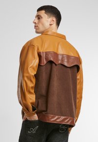 Braune und beige farbblockierte Jacke mit Leder- und Cordura-Texturen. Verfügt über ein gewelltes Leder-Backpanel mit geprägtem Schriftzug.