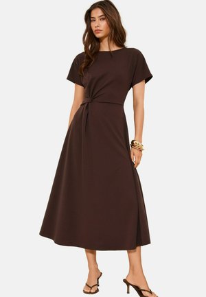 Frau trägt ein dunkelbraunes Midikleid mit kurzen Ärmeln und seitlichem Schlitz, kombiniert mit goldenen Armreifen und schwarzen Sandalen mit Absatz.