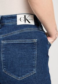 Jeans de mezclilla azul oscuro con una cintura alta, mostrando un bolsillo trasero y una etiqueta de tela blanca visible etiquetada como "Calvin Klein Jeans."