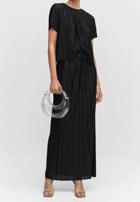 Schwarzes, plissiertes Maxikleid mit kurzen Ärmeln und elastischem Taillenbund. Kombiniert mit einer klaren, geschwungenen Handtasche und silbernen Sandalen mit Riemchen.