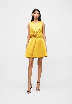 Γυναίκα που στέκεται με φόρεμα χωρίς μανίκια από κίτρινο σατέν με στριφτό κόμπο και λεπτομέρεια με ζώνη, φοράει μαύρα σανδάλια με ψηλά τακούνια.