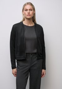 Svart kort jacka i slät tyg, med rund halsringning och dragkedja. Bärs över en mörkgrå skjorta och texturerade mörka byxor.