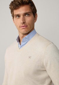 Suéter beige con escote en V, de textura de punto fino y con un pequeño logotipo bordado en el pecho. Llevado sobre una camisa a rayas de color azul claro.