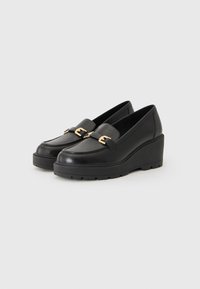 Mocassins compensés en cuir noir avec un bout arrondi, présentant un accent de boucle dorée et une semelle en caoutchouc texturée pour une meilleure adhérence.