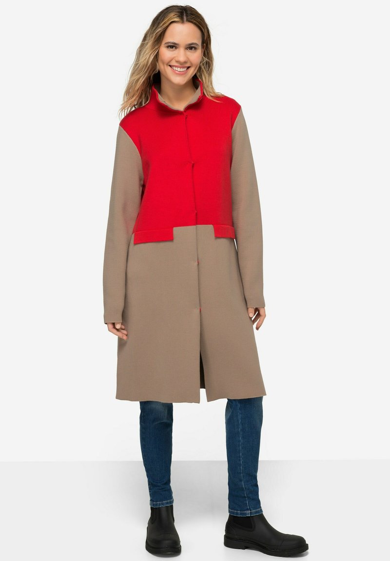 Cappotto lungo con una parte superiore rossa e una parte inferiore beige, realizzato in tessuto texturizzato con chiusura a bottoni e colletto.