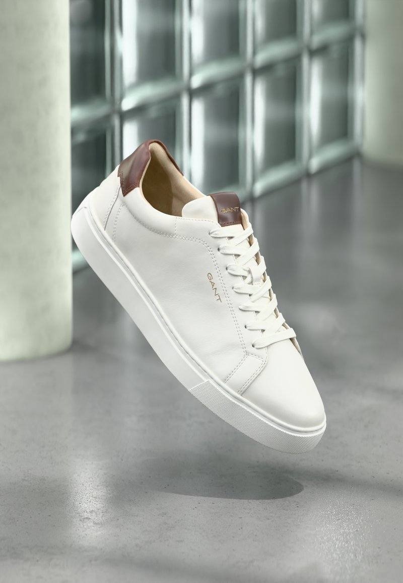 Vita lädersneakers med en hältab i brunt läder, snörningsdesign, gummisula och präglar logotyp på sidan. Sllät yta, minimalistisk stil.