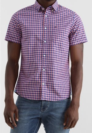 Man wearing short-sleeve button-up shirt with pink and blue checkered pattern and blue jeans against plain background.