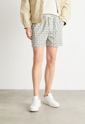 Personne portant une veste beige, un short à cordon de serrage à motifs, et des baskets blanches, debout sur un sol gris contre un mur blanc uni.