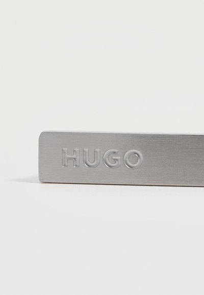 Праваправен метален правоъгълен етикет с издавена думата "HUGO" с главни букви на обикновен бял фон.