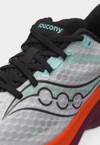 Κοντινή λήψη σε ένα παπούτσι τρεξίματος Saucony με μαύρα κορδόνια, λεπτομέρειες ανοιχτού τιρκουάζ, λευκό επάνω μέρος από δίχτυ και κόκκινο-πορτοκαλί άκρη στη σόλα.
