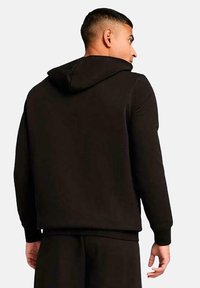 Sudadera con capucha negra hecha de una tela suave, con un diseño en la parte posterior, puños acanalados y un bolsillo tipo canguro, todo en un ajuste relajado.