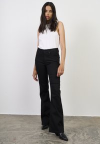 Femeie cu păr lung și închis la culoare, purtând un top alb fără mâneci, blugi evazați negri cu talie înaltă și cizme negre cu vârf ascuțit, stând în interior.