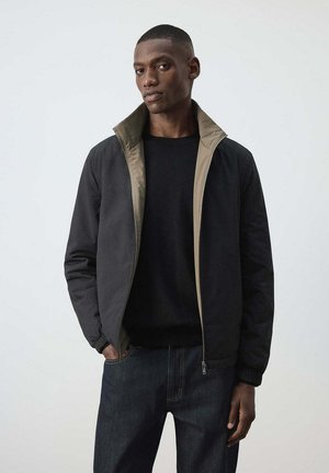 Hombre con chaqueta negra con cremallera forrada en beige y camisa negra, de pie con una mano en el bolsillo contra un fondo claro y liso.