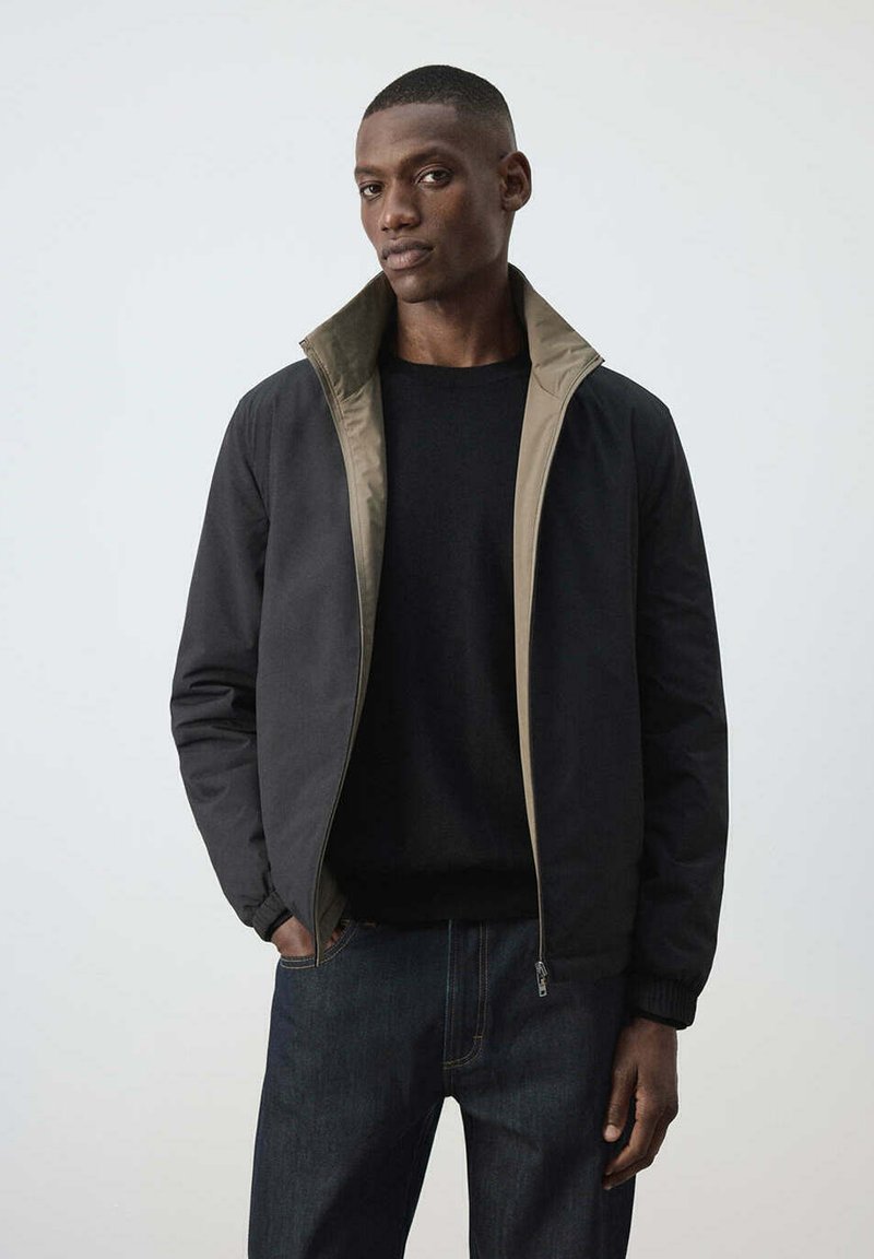 Homme portant une veste noire zippée avec une doublure intérieure beige et une chemise noire, debout avec une main dans la poche devant un fond uni clair.