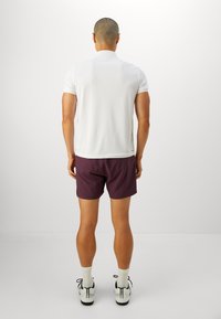 Bijela polo majica, kratkih rukava. Ljubičaste hlače do sredine butina. Uparene s bijelim čarapama i crno-bijelim tenisicama. Jednostavna pozadina.