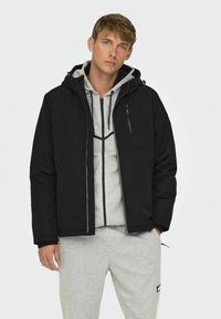 Czarna pikowana kurtka z odpinanym kapturem, zapinana na zamek błyskawiczny z przodu oraz z dwiema bocznymi kieszeniami. Noszona na jasnoszary hoodie oraz dresy.