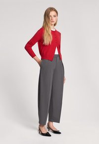 Cardigan rosso a maglia, camicia bianca, pantaloni larghi grigi e scarpe slingback con punta nera, in piedi contro uno sfondo neutro.