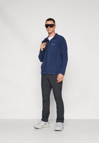 Granatowy polar z półsuwnym zamkiem i kieszenią na piersi, w połączeniu z ciemnymi spodniami i jasnoszarymi butami sportowymi. Minimalistyczny design i konstrukcja.