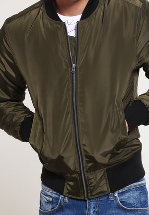 Mann trägt eine olivgrüne Bomberjacke mit Frontreißverschluss, schwarzen Bündchen und blauen Jeans, Hände in den Taschen.