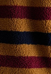 Fleece audums ar horizontālām svītrām sinepju dzeltenā, burgundijas un tumši zilā krāsā. Tā tekstūra ir mīksta un pūkaina, kas palielina komfortu un siltumu.